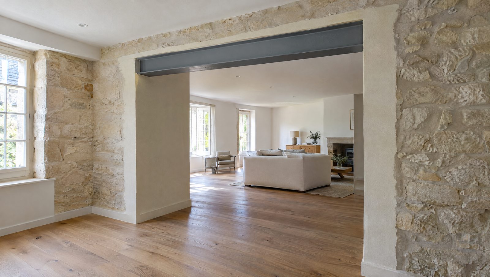 Large ouverture entre deux pièces d'une maison ancienne aux murs de pierre calcaire, poutre en acier peinte en gris visible dans le haut de la baie, tableaux fraîchement enduits et parquet de chêne, salon lumineux au fond
