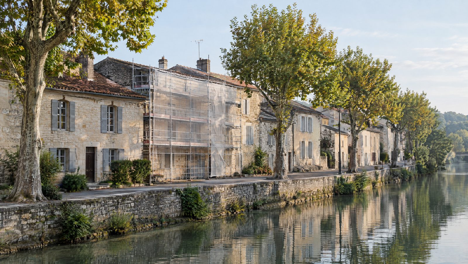 Maisons anciennes en pierre calcaire alignées le long d'un quai planté de platanes au bord d'une rivière, l'une d'elles sous un échafaudage bâché