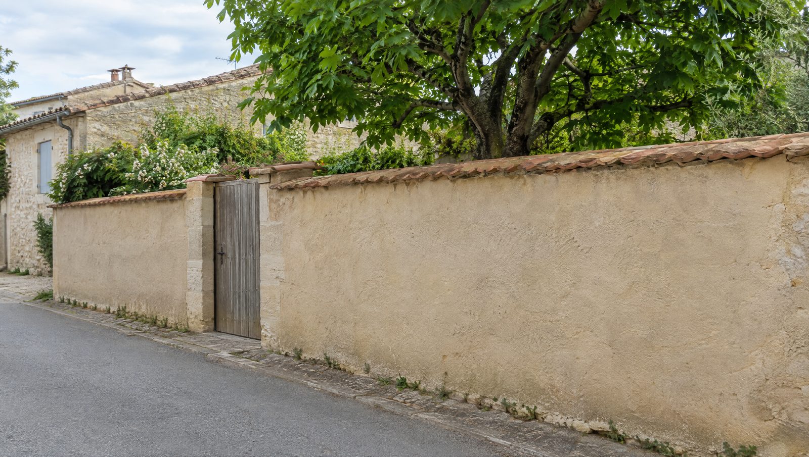 Mur de clôture enduit couleur sable et couronné de tuiles plates, avec un portillon en bois, le long d'une rue de petite ville
