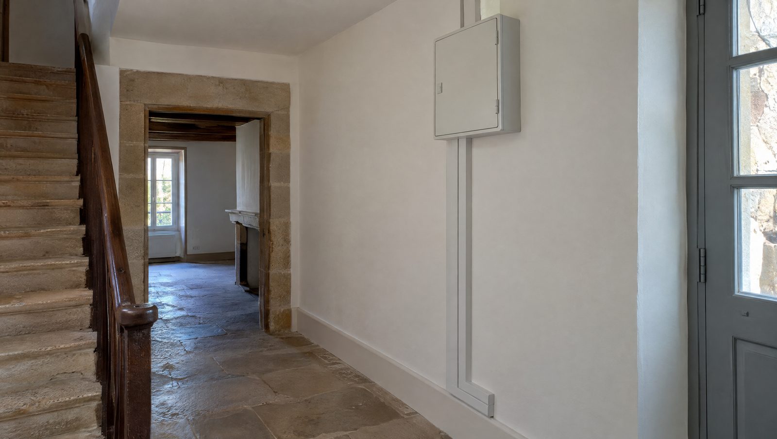 Tableau électrique gris fermé, fixé en hauteur dans l'entrée d'une maison ancienne et alimenté par une goulotte le long du mur blanc, près d'un escalier en bois, d'un sol en dalles de pierre et d'un encadrement de porte en pierre