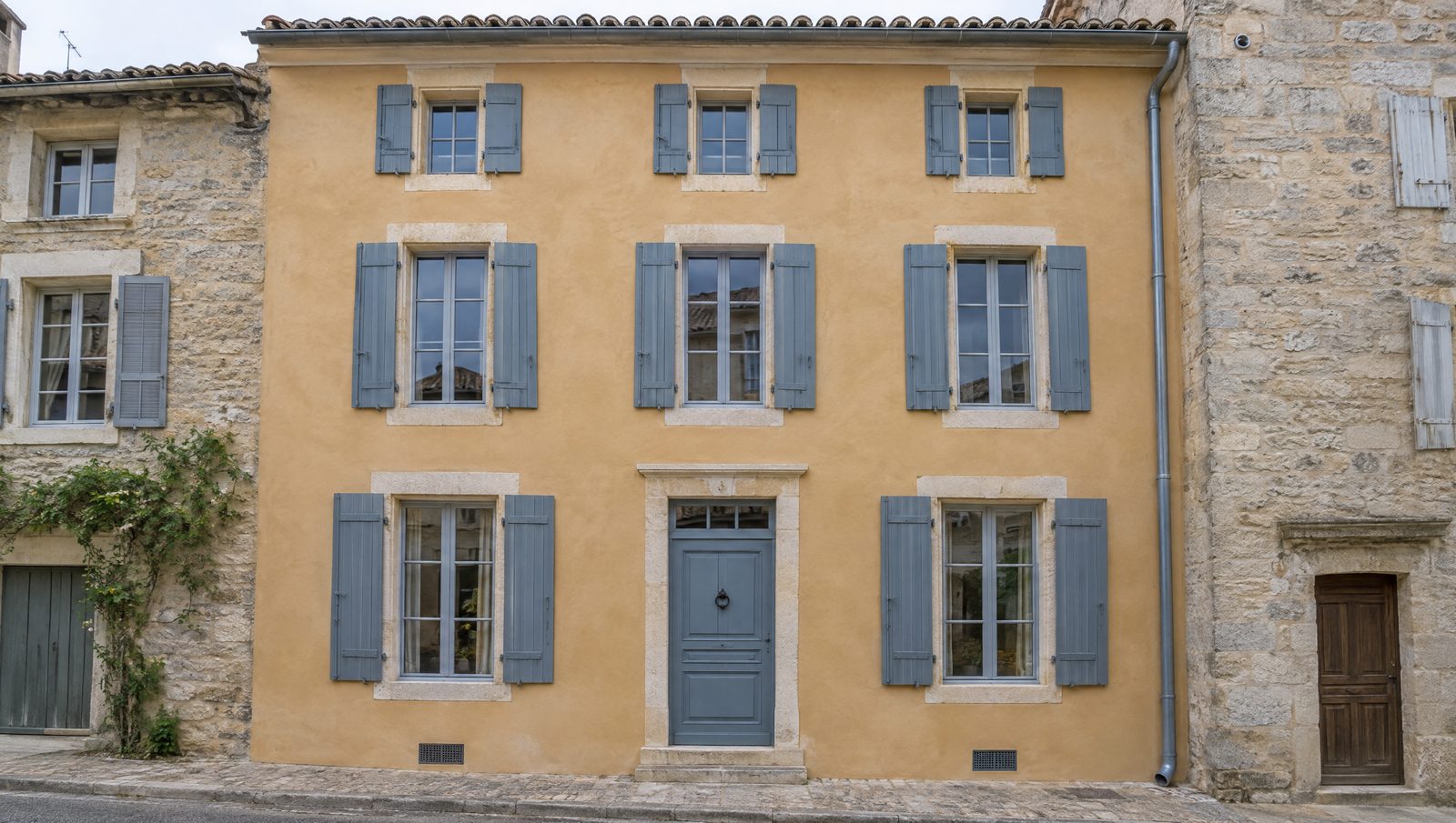 Façade fraîchement restaurée d'une maison de ville ancienne, enduit à la chaux ocre sable, encadrements en pierre calcaire, volets en bois gris bleu et descente en zinc, entre deux maisons de pierre