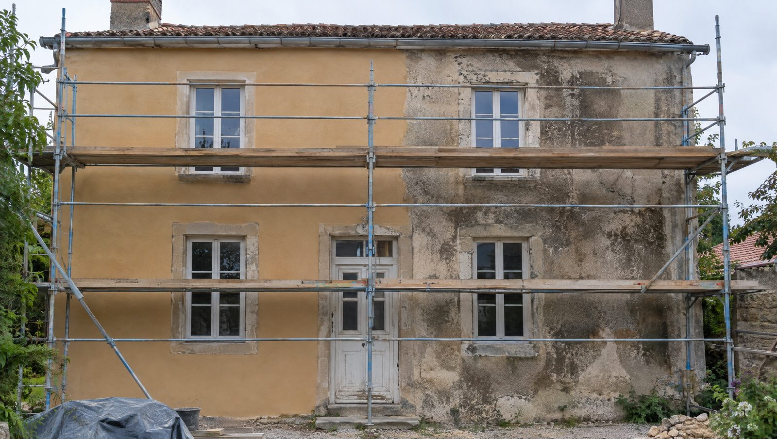 Façade d'une maison ancienne à deux niveaux sous un échafaudage en acier, une moitié du mur couverte d'un enduit à la chaux ocre sable neuf, l'autre encore tachée, volets déposés
