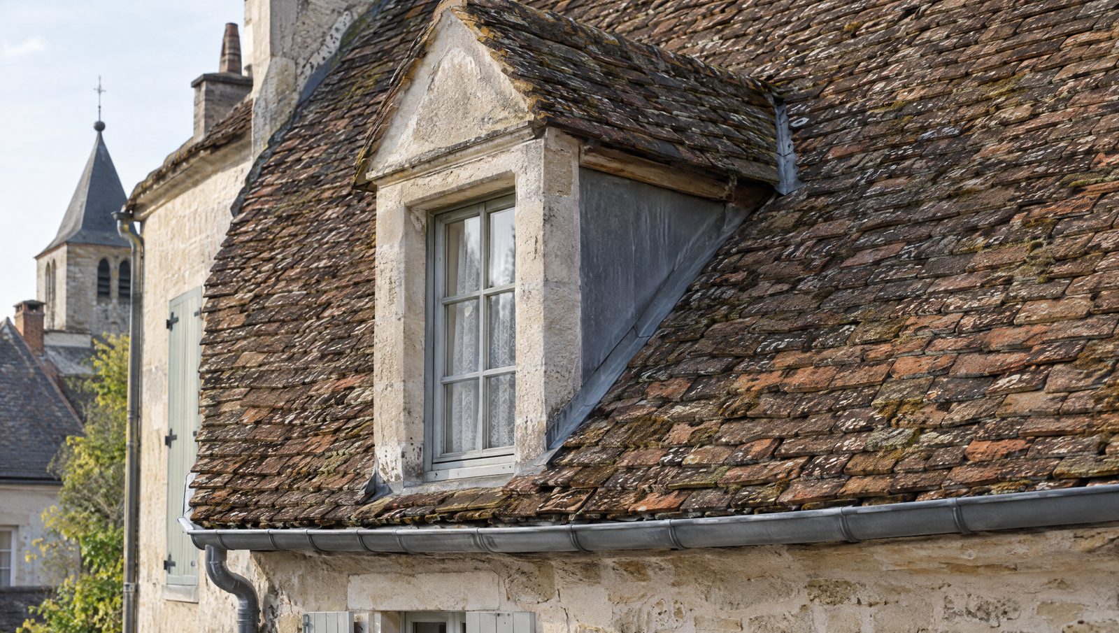 Toit ancien en petites tuiles plates de terre cuite vieillies, avec une lucarne en pierre et une gouttière en zinc, sur une maison en pierre calcaire d'un bourg historique