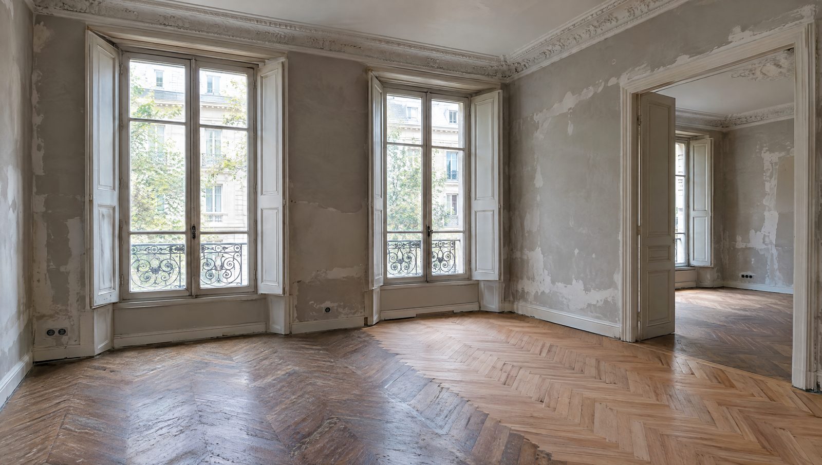 Appartement vide d'une maison de ville ancienne en travaux, hautes fenêtres à volets intérieurs, moulures au plafond, murs en cours d'enduit et parquet à bâtons rompus en partie poncé