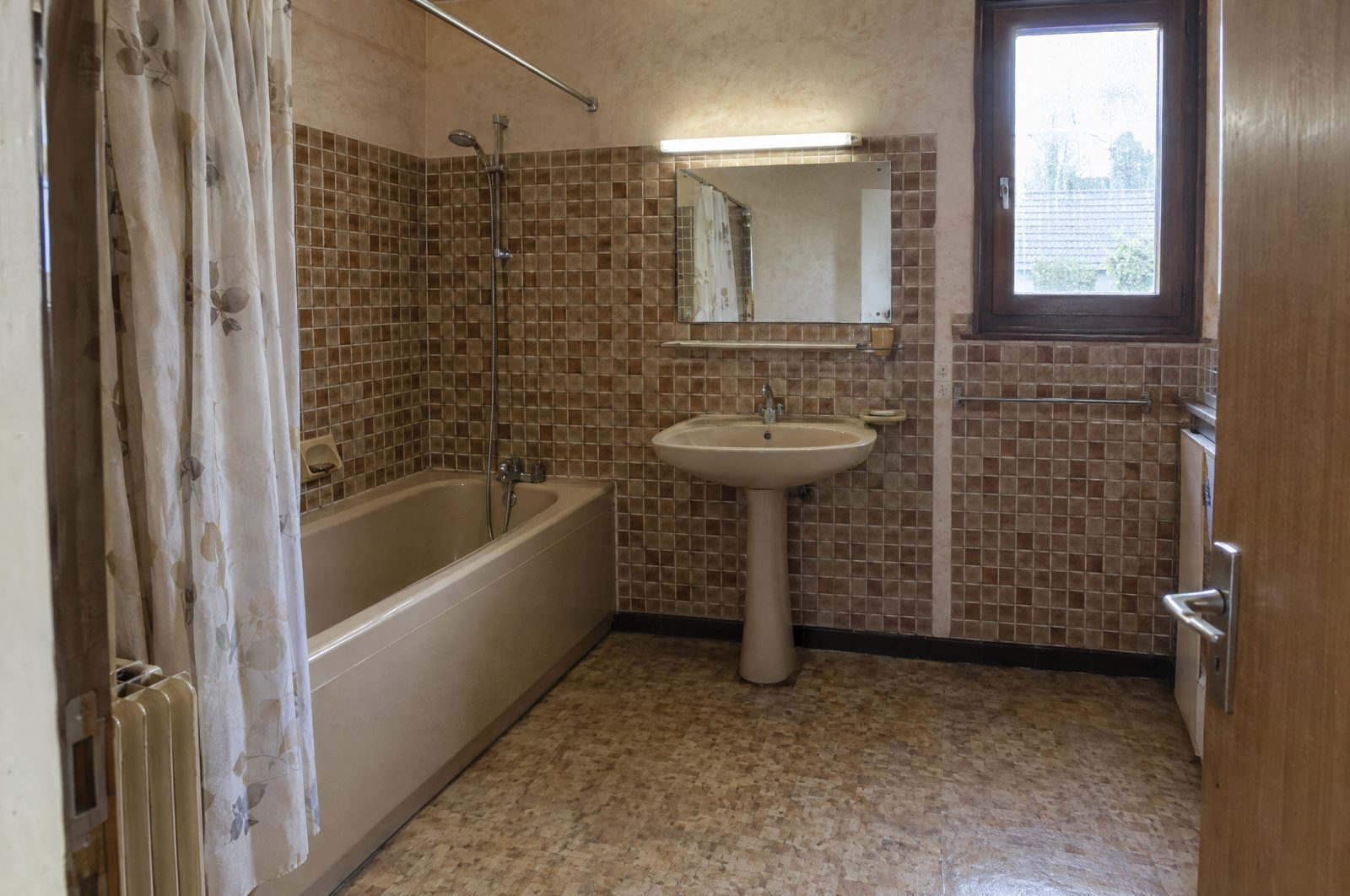 Salle de bain des années 1980 avant rénovation, baignoire beige avec rideau de douche à fleurs, petite mosaïque brune et beige, lavabo sur colonne et sol en linoléum