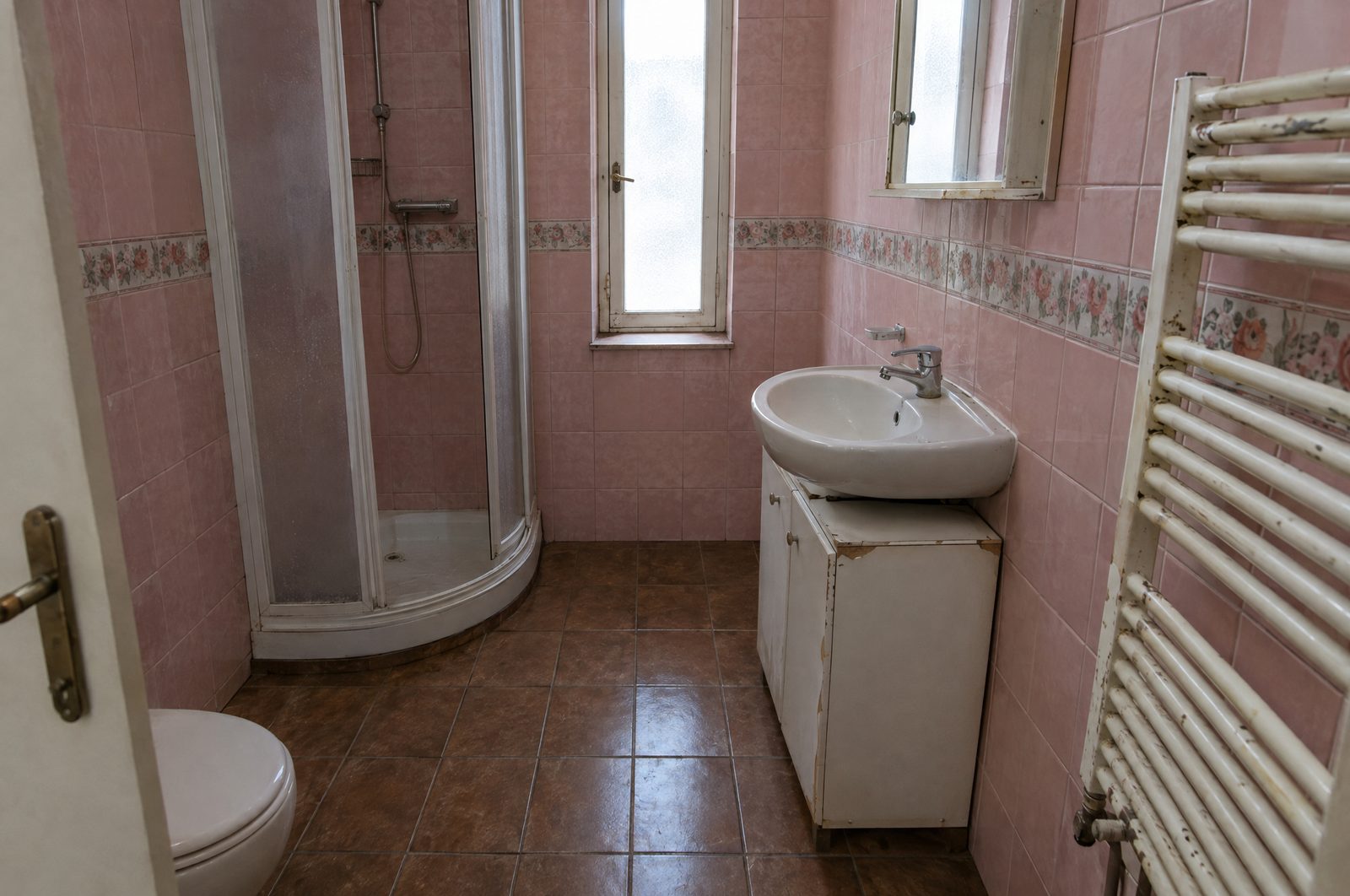 Petite salle de bain d'appartement avant rénovation, faïence rose à frise fleurie, cabine de douche d'angle, lavabo sur un meuble blanc écaillé, vieux sèche-serviettes et sol en carreaux bruns