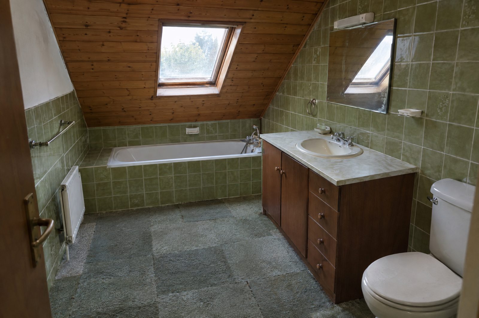 Salle de bain sous combles avant rénovation, rampant en lambris verni, baignoire sous une fenêtre de toit, faïence verte, meuble vasque en bois foncé et sol en dalles de moquette