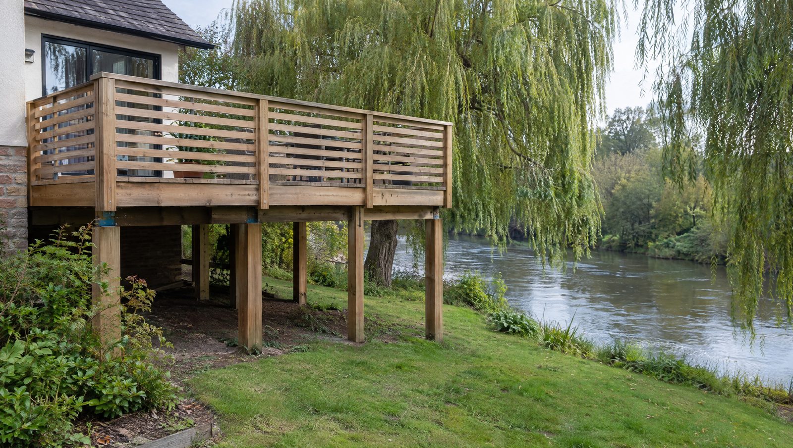 Terrasse en bois surélevée sur poteaux en bois à l'arrière d'une maison, avec un garde-corps à claire-voie et un vide laissé ouvert sous le plancher, devant une rivière calme bordée de saules